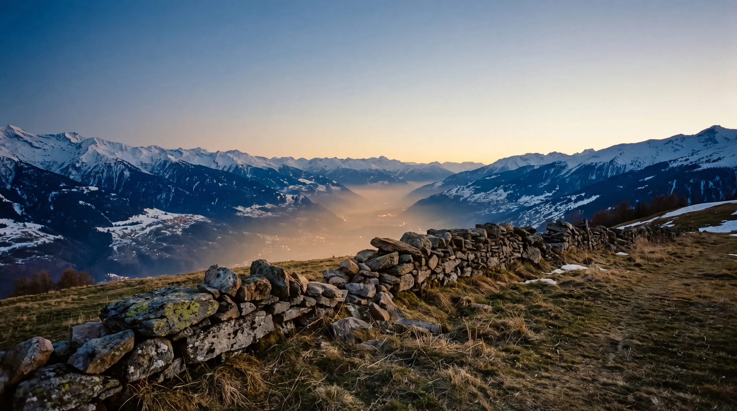 Une vallée de montagne à la tombée du jour, vue depuis un pâturage en hauteur : un muret de pierres sèches court au premier plan, les lumières d'une agglomération ne forment qu'une lueur ambrée au fond de la vallée noyée de brume, et des crêtes enneigées se referment de part et d'autre sous un ciel dégradé sans nuage.